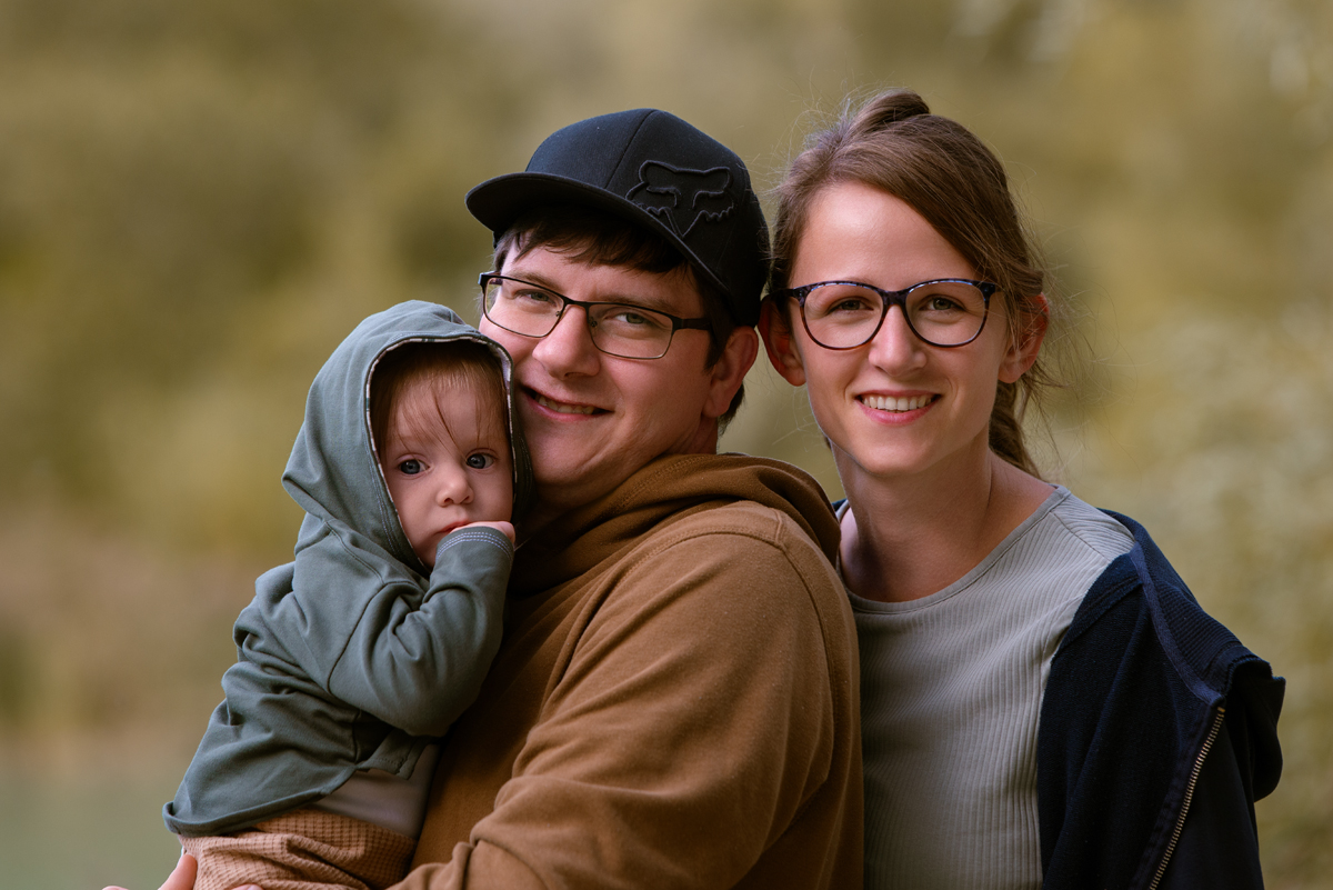 Junges Paar steht eng beisammen und hält ihr Baby im Arm, alle drei lächeln in die Kamera vor einem weichen, natürlichen Hintergrund.